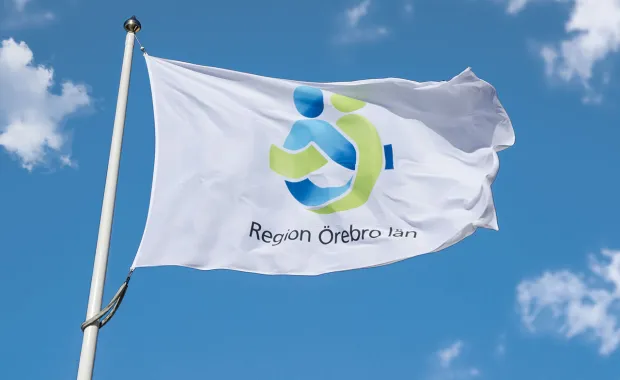 En vit flagga med logotypen för Region Örebro län vajar i vinden mot en klarblå himmel med några moln.