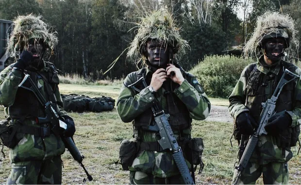 Tre militärklädda män med hjälmar och vapen samt målade i ansiktet står och tittar mot kameran ute på fältet