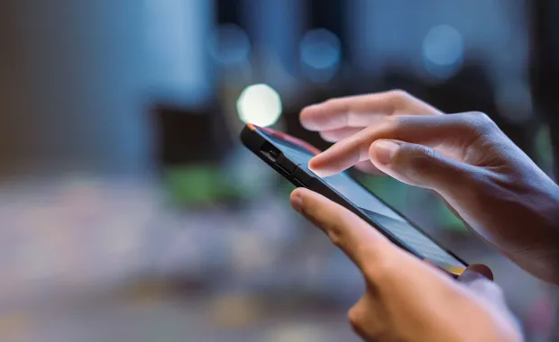 Närbild av en hand som använder en smartphone med pekskärm. Fingret trycker på skärmen i en mörk, suddig miljö som antyder digital användning.