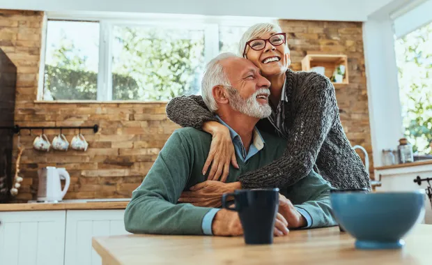 Ett äldre par sitter vid ett köksbord i ett ljust och hemtrevligt kök. Kvinnan kramar mannen bakifrån medan båda skrattar och ser lyckliga ut. Kaffekoppar och en skål står på bordet.