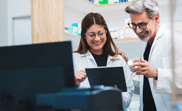 Två vårdmedarbetare i vita arbetskläder står tillsammans och granskar information på en surfplatta. De ler och diskuterar innehållet i en modern vård- eller laboratoriemiljö.