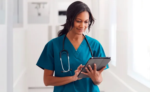Kvinnlig läkare i blågröna sjukhuskläder och stetoskop runt halsen står och tittar på sin surfplatta