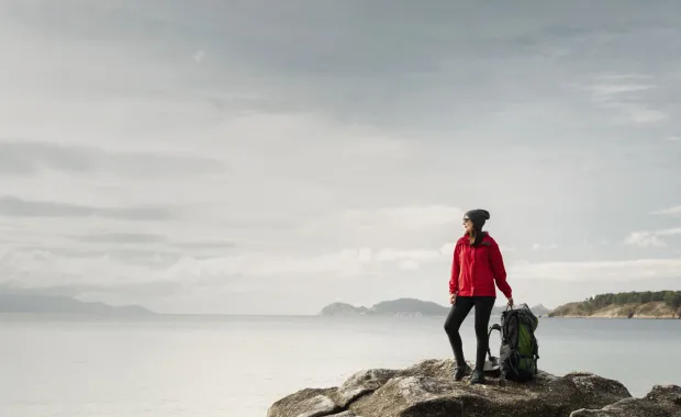 Kvinna i röd jacka och mössa står med en stor ryggsäck bredvid sig på en klippa vid havet och blickar ut