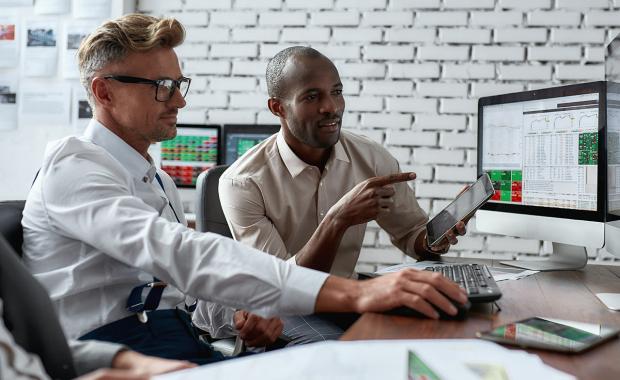 Två manliga kollegor sitter tillsammans vid ett bord framför stora dataskärmar med finansiell data och grafer