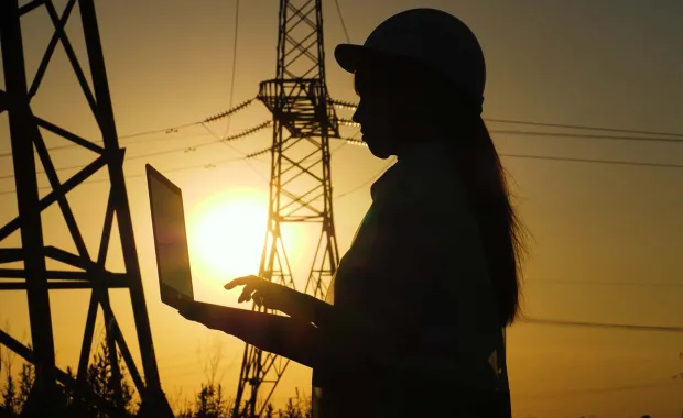 a silhouetted electrical engineer records data on a laptop next to electrical towers at sunset