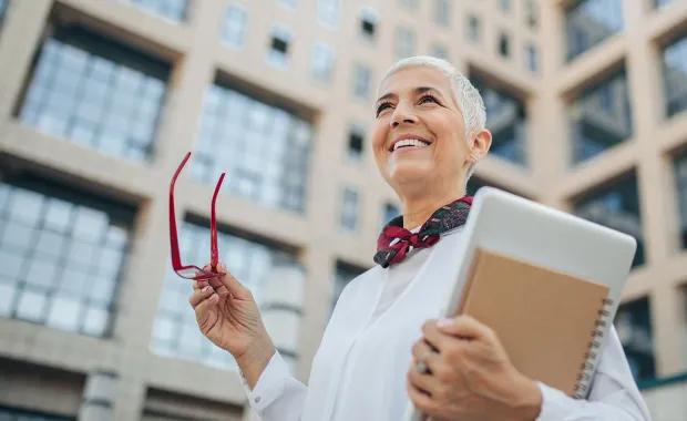 Kvinna står leende utanför kontoret med dokument i ena handen och sina glasögon i den andra