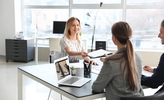 Kvinna sträcker leende över dokumentation till annan kvinna vid ett mötesbord på ett stort och ljust kontor