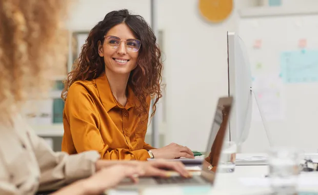 Kvinna med glasögon tittar leende upp från sin laptop för att lyssna på kollega