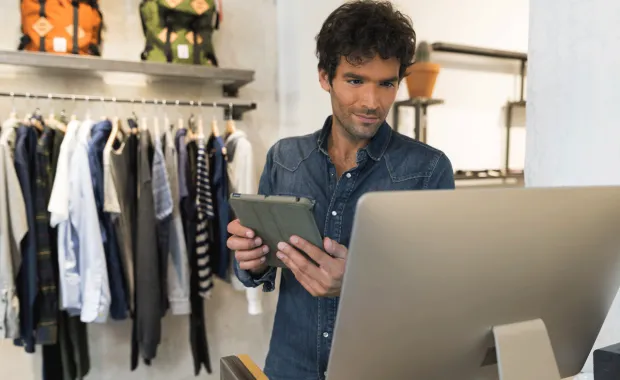 Manlig expedit med surfplatta står i en klädesbutik och tittar på dataskärmen i kassan