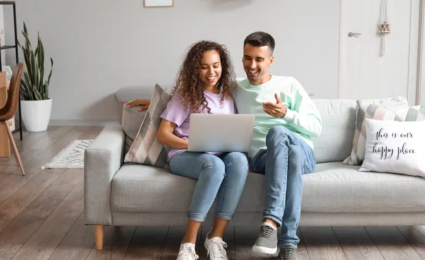 A happy looking couple sitting on their sofa, smiling while looking at a laptop