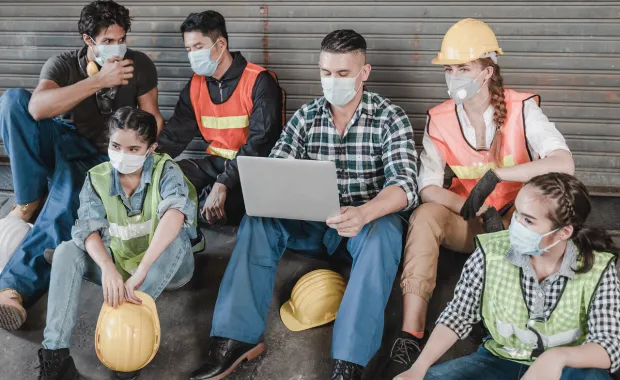 group of factory workers in PPE sit together around a laptop