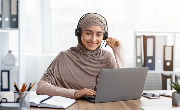 smiling lady wearing a hijab and headset working on a laptop