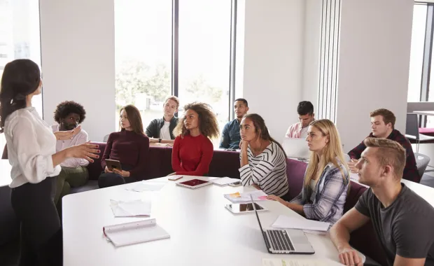A presenter stands at the front of a meeting room, speaking to a diverse group of students or professionals
