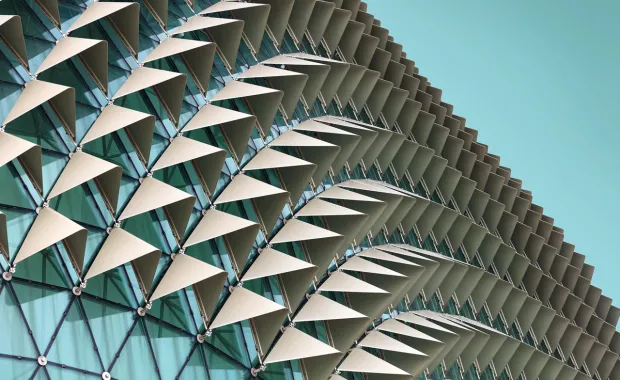 Close-up of a modern glass building facade with rows of white triangular sunshades arranged in a repeating geometric pattern against a clear blue sky.