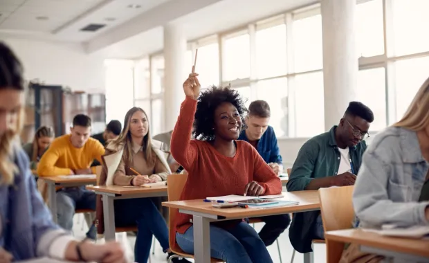 A smiling student raises her hand during a lecture as classmates take notes