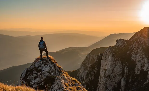 En person med ryggsäck står på en bergstopp och blickar ut över ett bergslandskap i soluppgångens gyllene ljus. Utsikten visar flera lager av berg i dimma.