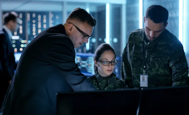 Kvinnlig militär sitter vid datorn och två manliga kollegor står bakom och tillsammans tittar de på skärmen