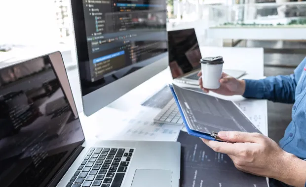 En person sitter med en kaffemugg i ena handen och läser på sin surfplatta framför dataskärmar på skrivbordet