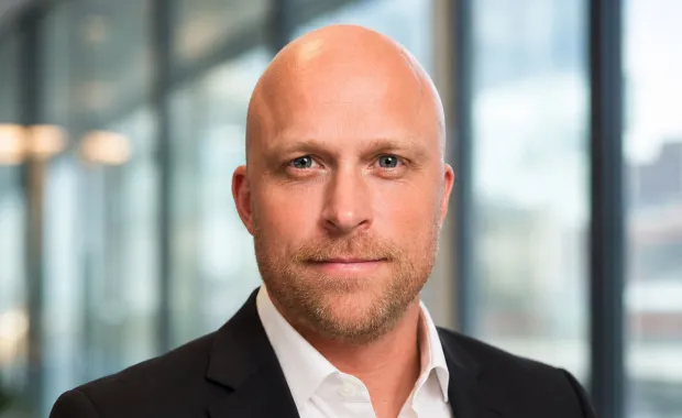 Porträtt av en man i kostym och vit skjorta som ser in i kameran. Bakgrunden är mjukt suddig med stora fönster och en modern kontorsmiljö, vilket ger ett professionellt och förtroendeingivande intryck.