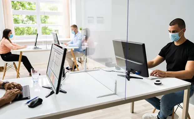 Man med munskydd sitter och jobbar vid sin laptop i ett öppet och ljust kontorslandskap där två kvinnliga kollegor syns i bakgrunden