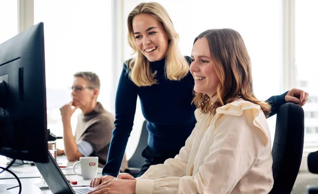 To personer smiler foran dataskjerm, tredje sitter i bakgrunnen.