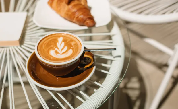 En kopp latte med skummönster står på ett cafébord tillsammans med en croissant på en tallrik. Bordet är av glas med vit metallstomme och miljön är ljus och inbjudande.