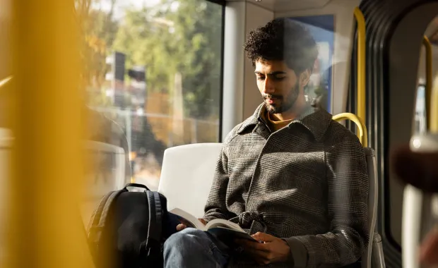 En man sitter vid fönstret i en buss eller spårvagn och läser en bok. Solljus faller in genom fönstret och en ryggsäck står bredvid honom.