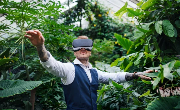 Man wearing VR headset reaching arms out against background of green leaves