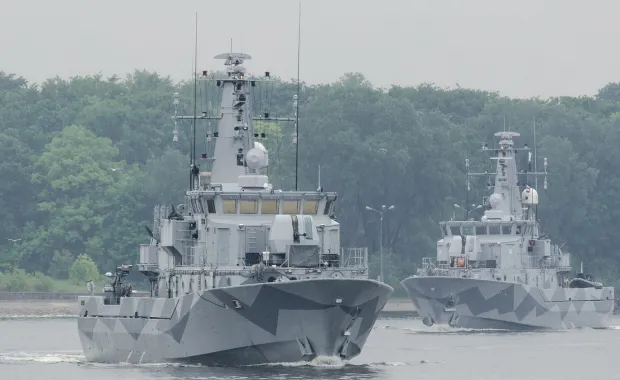 Två militärfartyg på öppet hav med skog och land i bakgrunden