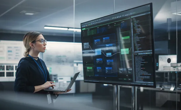 Woman holding a laptop and looking at a large monitor