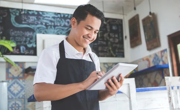 Male café owner wearing a black apron smiling while using a tablet