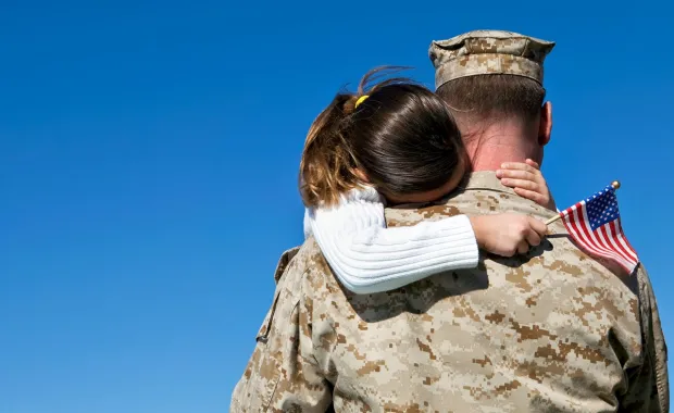 Military man standing and hugging daughter who is holding an American flag