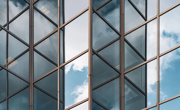 Närbild av en modern byggnads glasfasad med geometriska metallramar. Blå himmel och moln speglas i glasrutorna och skapar ett abstrakt arkitektoniskt mönster.