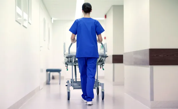 Kvinna i blåa sjukhuskläder går i en sjukhuskorridor med en patientsäng
