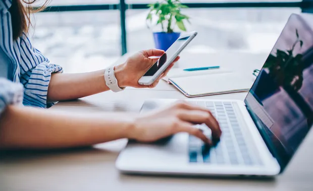 Närbild av en person som arbetar vid en laptop samtidigt som personen håller en smartphone. På skrivbordet ligger anteckningsmaterial, vilket illustrerar digitalt arbete och användning av flera enheter.