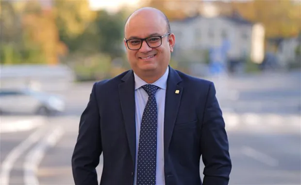 Porträtt av en leende man i kostym och slips, stående utomhus i en stadsmiljö med suddig bakgrund i höstfärger. Personen bär glasögon och ser professionell ut.