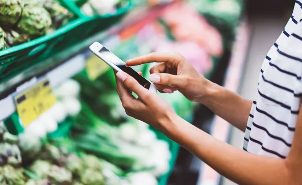Frau steht im Supermarkt in der Obst- und Gemüseabteilung und schaut auf ihr Handy