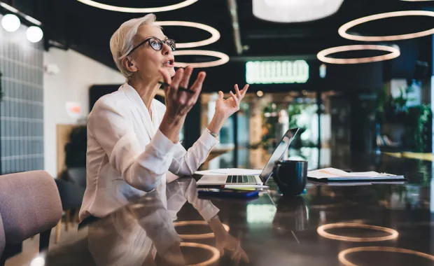 En kvinna med glasögon sitter vid ett bord i ett modernt kontor och deltar i ett videosamtal via laptop.