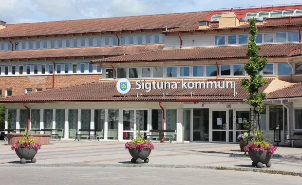 Entrén till Sigtuna kommuns kommunhus med skylten "Sigtuna kommun" ovanför ingången. Byggnaden har tegel- och putsfasad med röda takpannor, och framför entrén står stora blomkrukor med färgglada blommor.