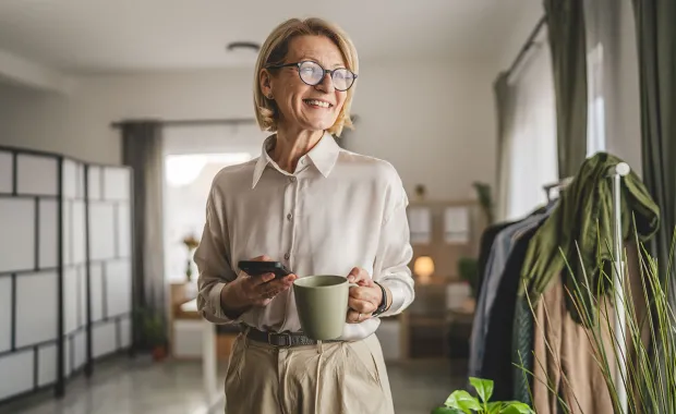 Äldre kvinna med glasögon står inomhus, håller en kopp kaffe och en mobiltelefon, och ler.