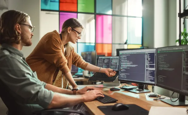 Två mjukvaruutvecklare samarbetar vid flera datorskärmar fyllda med kod i ett modernt kontor. En kvinna pekar på skärmen medan en man skriver på tangentbordet. Bakgrunden visar färgade glaspartier som skapar en kreativ arbetsmiljö.