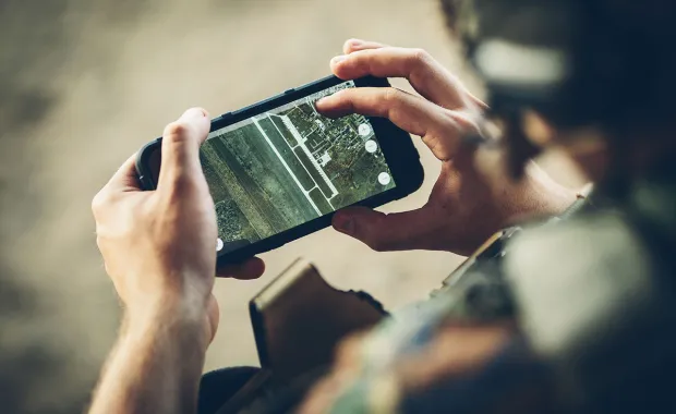 Närbild av en person i militäruniform som håller en smartphone med en digital karta eller satellitbild på skärmen. Bilden visar användning av mobil teknik för navigering, lägesbild eller informationshantering i fältmiljö.
