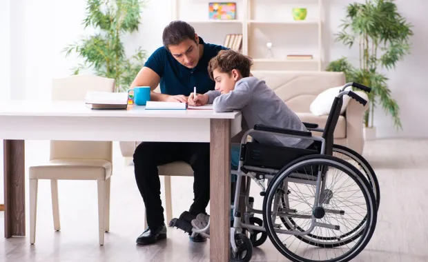 Adult helping a child in a wheelchair with schoolwork at a table in a bright, comfortable home setting