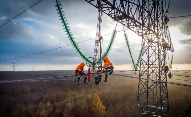 Två tekniker i orange skyddskläder arbetar högt upp på en kraftledningsstolpe med elledningar och isolatorer i solnedgång.
