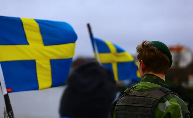 Svenska flaggor vajar i vinden medan en person i uniform står med ryggen mot kameran. Bilden förmedlar en ceremoniell eller patriotisk stämning i en utomhusmiljö.