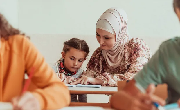 En kvinnlig lärare med slöja hjälper en ung elev med skolarbete i klassrummet. De sitter tätt tillsammans vid en bänk, fokuserade på skrivboken. Andra elever syns suddigt i förgrunden.