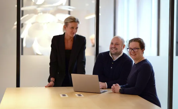 Tre kollegor står och sitter runt ett bord i ett modernt kontor och samarbetar kring en laptop. De ler och verkar engagerade i diskussionen.
