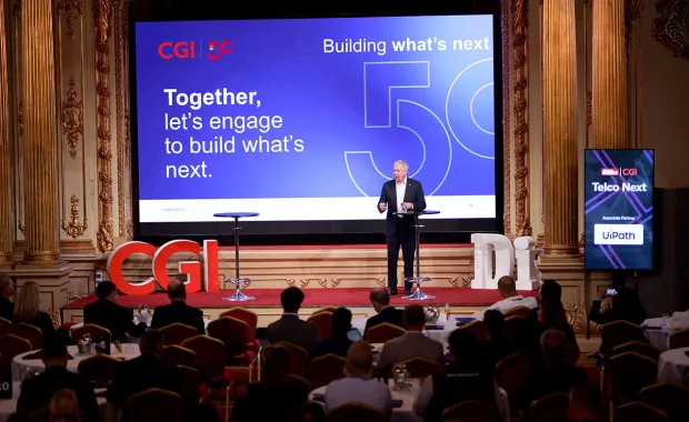 En talare står på scen under ett CGI-evenemang inför en sittande publik i en elegant konferenssal. På den stora skärmen bakom syns texten “Together, let’s engage to build what’s next” samt CGI:s 50-årsjubileumsgrafik.