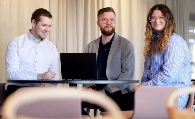 Tre kollegor sitter tillsammans vid ett bord med en laptop och samarbetar. De ler och verkar ha en avslappnad och positiv diskussion i en modern kontorsmiljö.