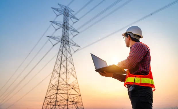 Arbeider med hjelm og refleksvest bruker laptop ved høyspentmast.